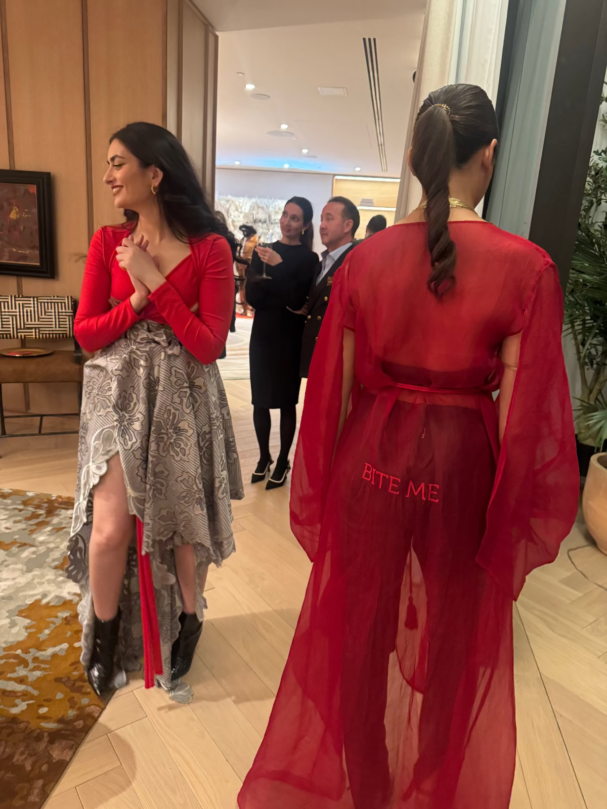 Two fashion models, one in a Priscilla Couture red outfit with 'BITE ME' statement and the other in a patterned grey skirt, in an indoor setting.