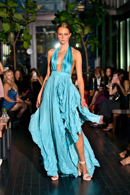 Model wearing a pool blue dress on a runway with an audience in the background.