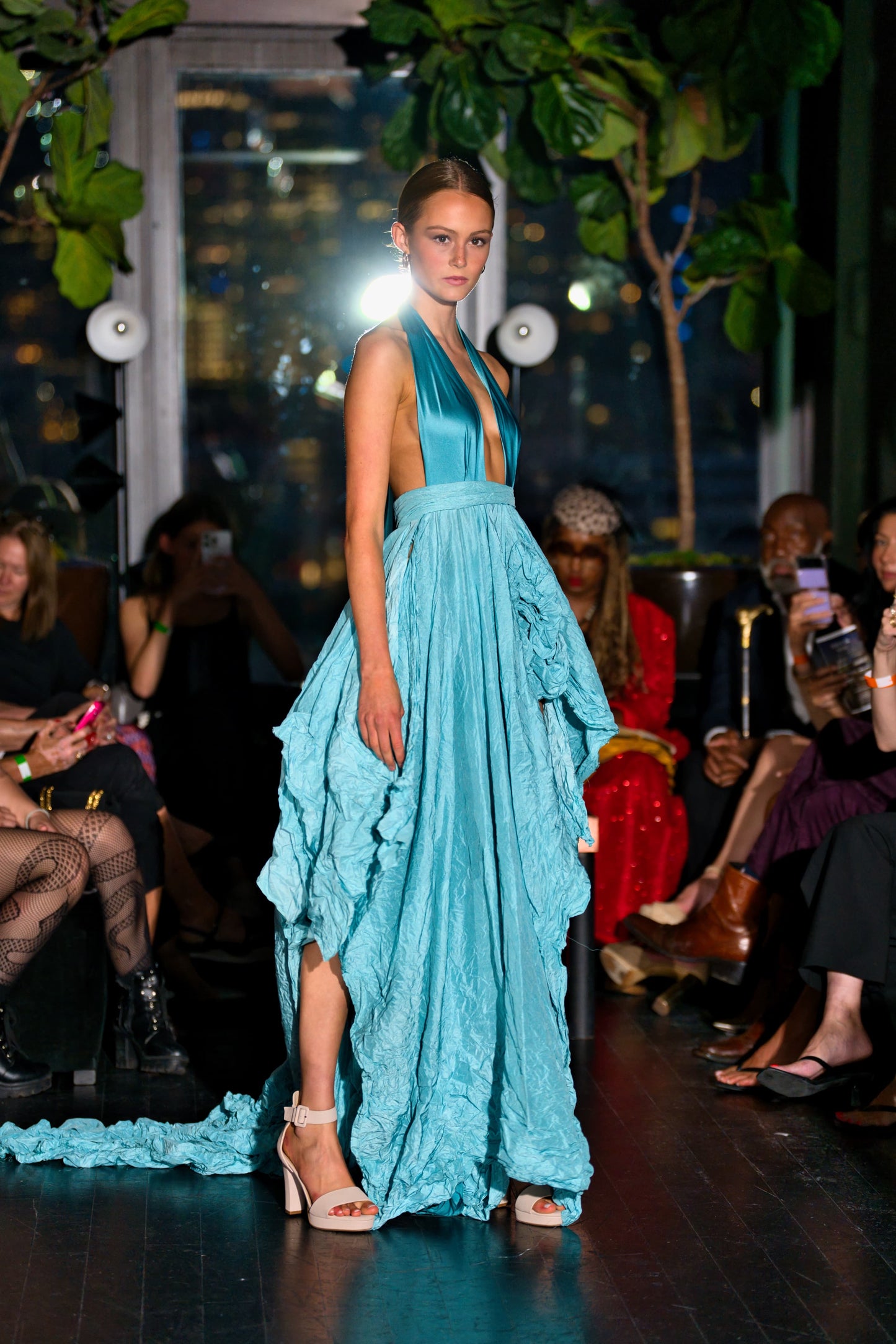 Model wearing a pool blue evening gown on a runway with audience members in the background.