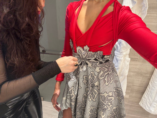 Fashion model wearing a Priscilla Couture red top and patterned grey skirt with another fashion designer adjusting something on the waist.