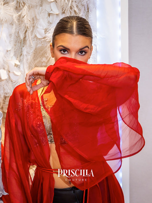 A model wearing a vibrant red sheer silk organza robe with dramatic wide sleeves over a gold embroidered bralette, posed against a white feathered wall. The brand name 'PRISCILLA COUTURE' is watermarked at the bottom.