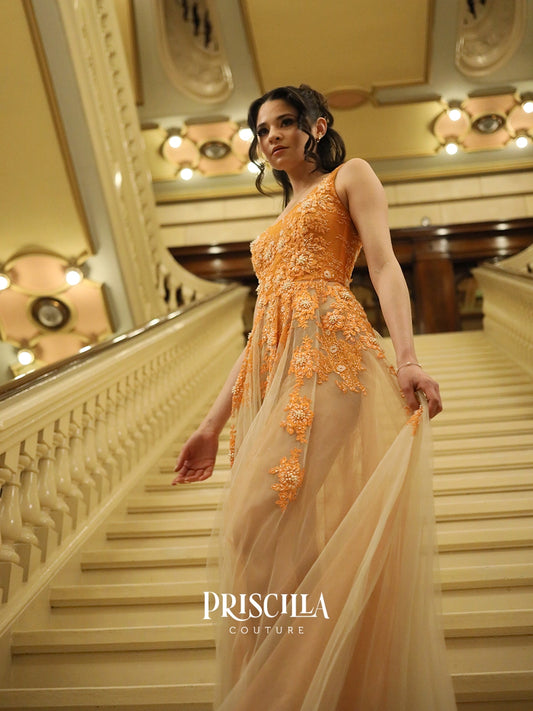 A Priscilla Couture fashion model wearing a Priscilla Couture sleeveless orange gown with intricate floral embroidery and sheer tulle, walking down a grand staircase in a luxurious interior. The brand name 'PRISCILLA COUTURE' is visible as a watermark at the bottom.