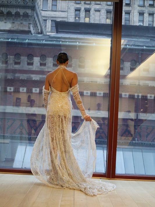 Woman in a gold evening gown standing in front of a large window with a cityscape reflection.
