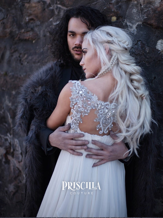 A woman in a white bridal gown with an ornate beaded back and long platinum-blonde hair is embraced by a man in a dark fur-lined cloak against a rocky background. The brand name PRISCILLA COUTURE is printed at the bottom.