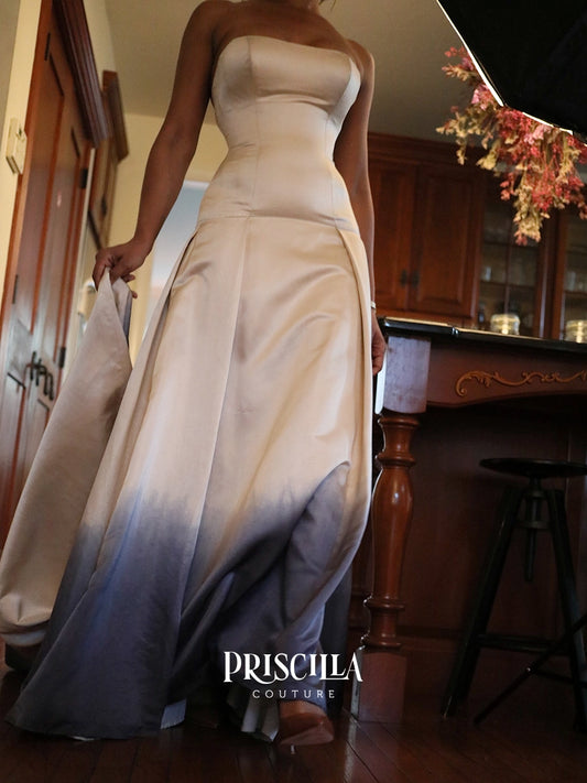 A strapless floor-length silk duchesse gown worn by a model in an indoor room with wood floors and cabinetry. The dress has a fitted bodice and a flowing skirt with a smooth gradient from warm ivory at the top to slate blue-gray at the bottom.