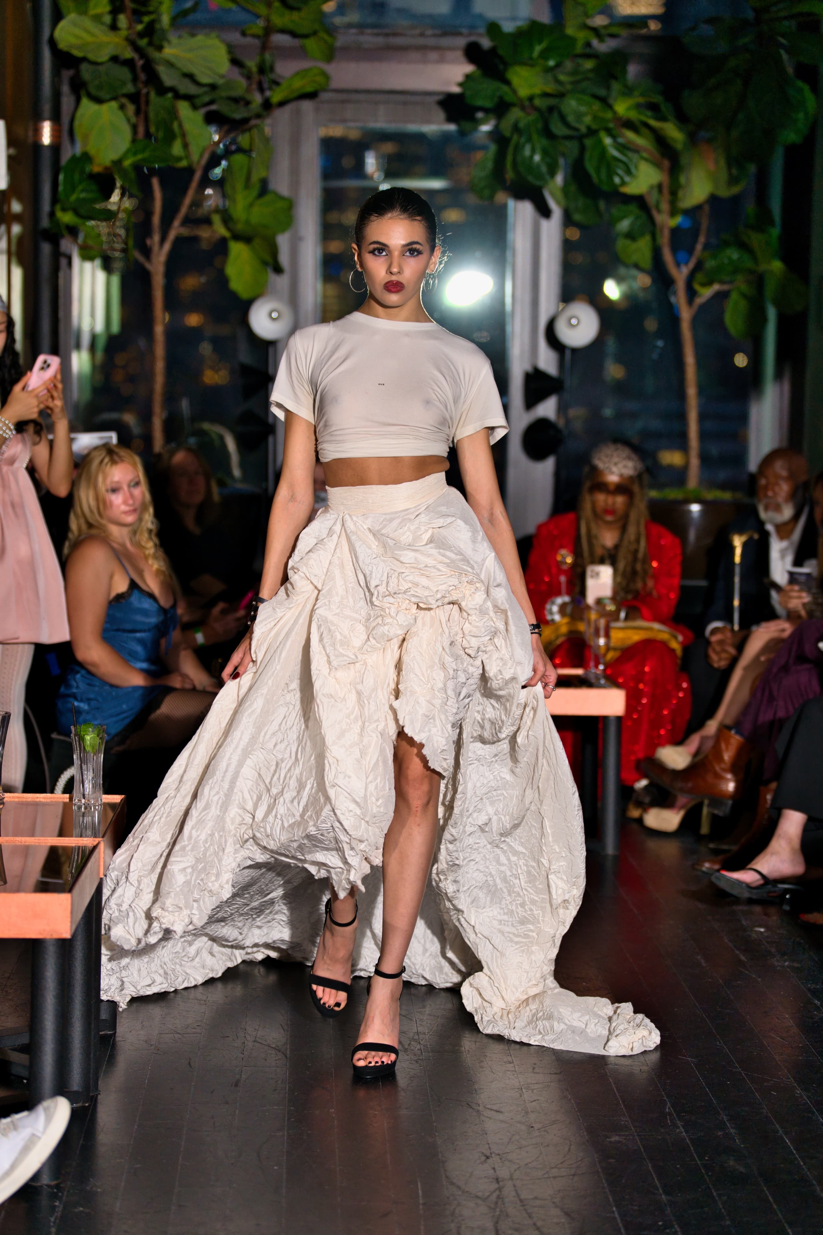 Model walking on a runway wearing a F white tee outfit with a rebellious, textured skirt.