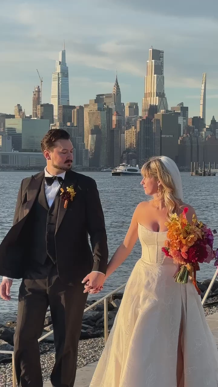 Bride in a Priscilla Couture custom wedding gown with her groom walking by the river. NYC skyline in the background.