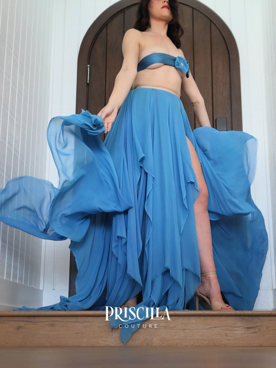 A model wearing a Priscilla Couture blue strapless bandeau top and matching high-slit Priscilla Couture maxi skirt, holding out the flowing chiffon skirt on wooden stairs in front of a white paneled wall and dark wood door.