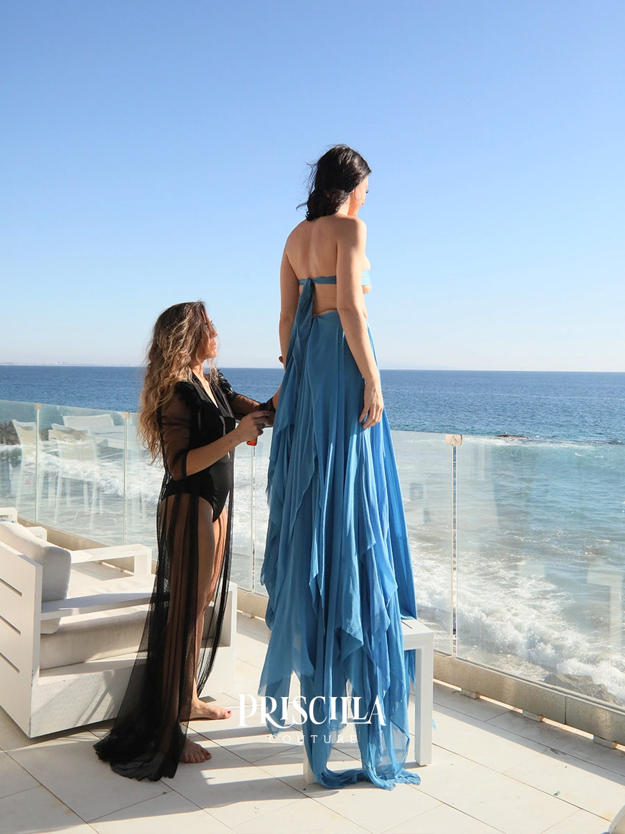 A woman wearing a Priscilla Couture layered blue maxi silk chiffon convertible skirt stands on a sunlit terrace by the ocean while fashion designer adjusts the skirt.