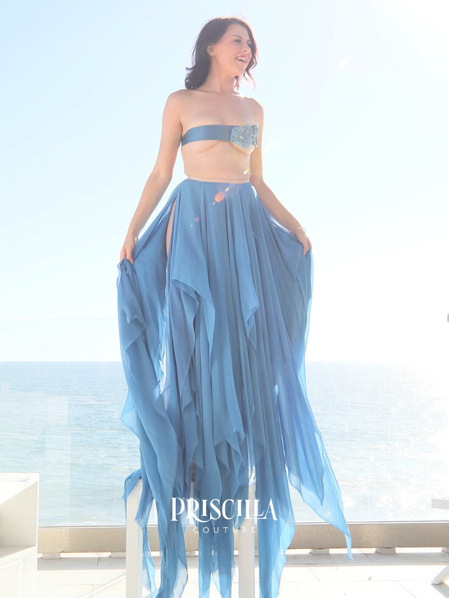 A model wearing a light blue two-piece Priscilla Couture set consisting of a strapless bandeau top with a floral applique at the center and a matching high-slit maxi skirt, standing outdoors with the ocean in the background.
