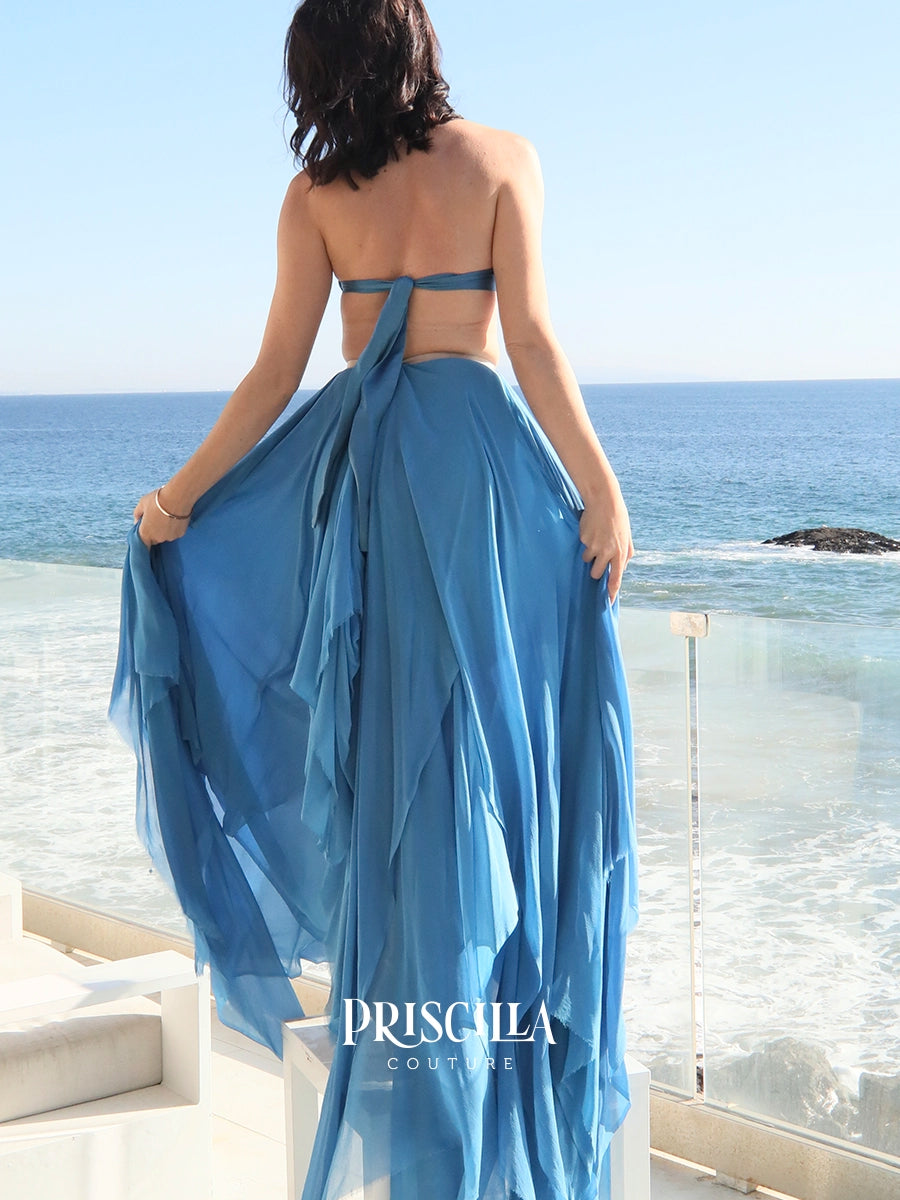 A woman stands with her back to the camera wearing a flowing Priscilla Couture blue skirt with a bralette, holding the skirt out over a beachfront balcony with the ocean in the background. 
