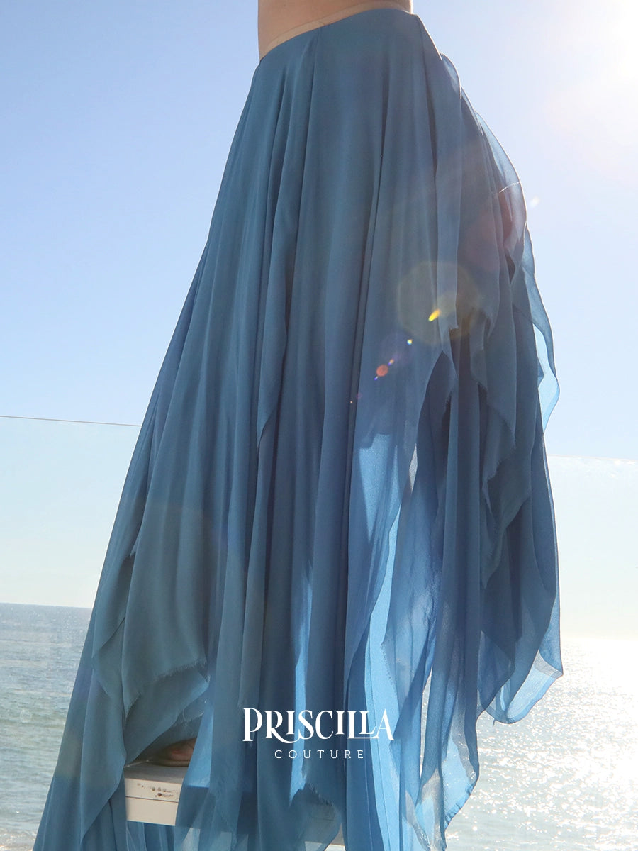 A flowing Priscilla Couture sheer blue maxi skirt worn outdoors by a model, with the ocean and bright sky in the background.