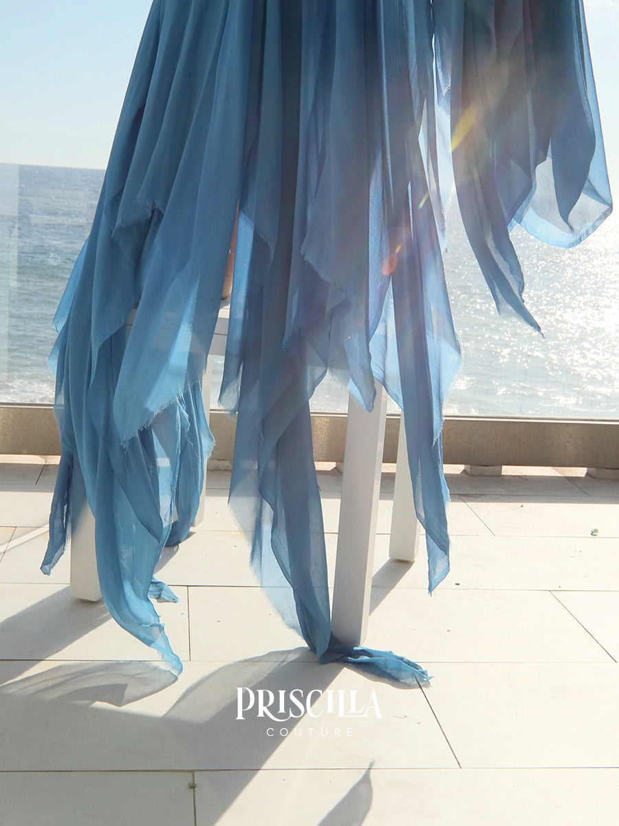A flowing Priscilla Couture sheer blue skirt fabric drape hanging from white support stands on a sunlit terrace with the ocean visible in the background. 