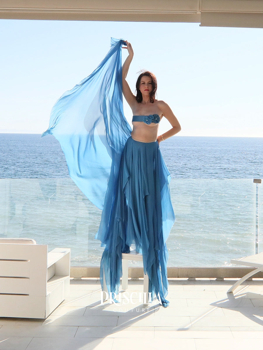 A woman modeling a two-piece Priscilla Couture blue bridal wear set consisting of a strapless bandeau top and a matching tiered maxi skirt, standing on a sunlit balcony with the ocean in the background.
