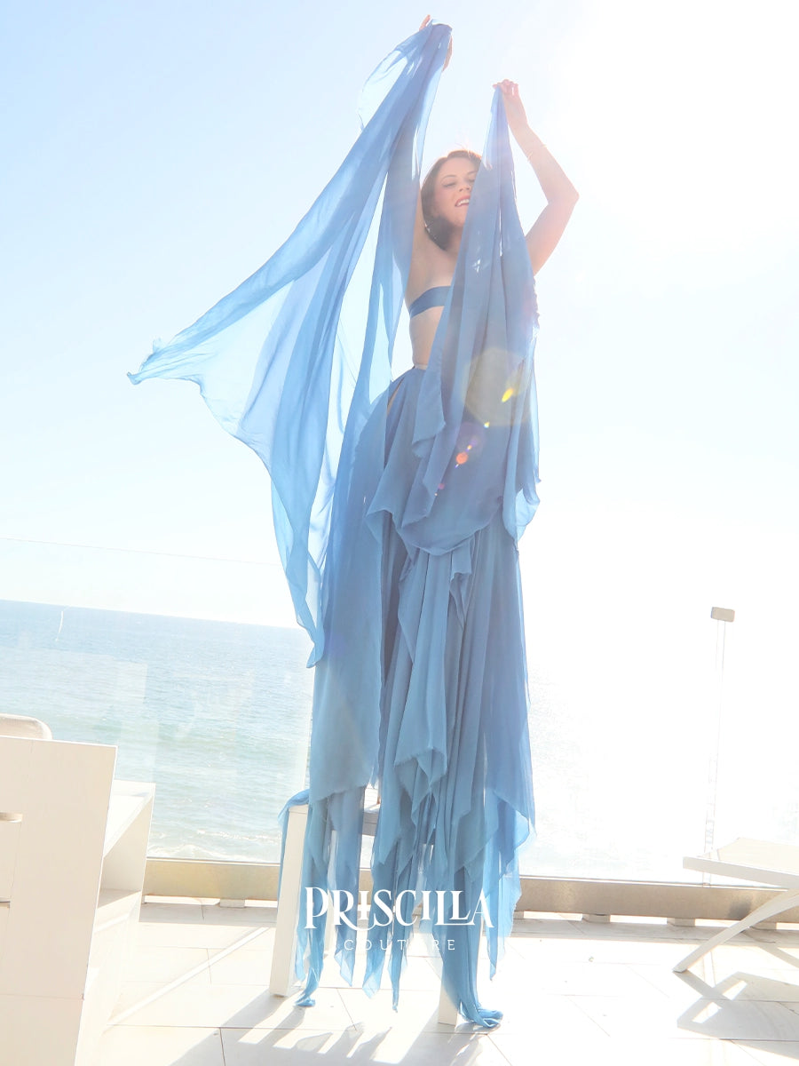 A woman modeling a flowing Priscilla Couture light blue sheer skirt, standing on a sunlit terrace with the ocean in the background. 