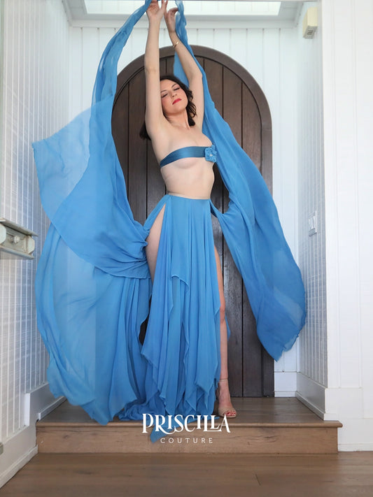 A woman modeling a Priscilla Couture  blue satin bandeau top and matching high-slit maxi skirt with flowing sheer blue fabric draped from her raised arms, posed indoors in front of a dark arched door. 