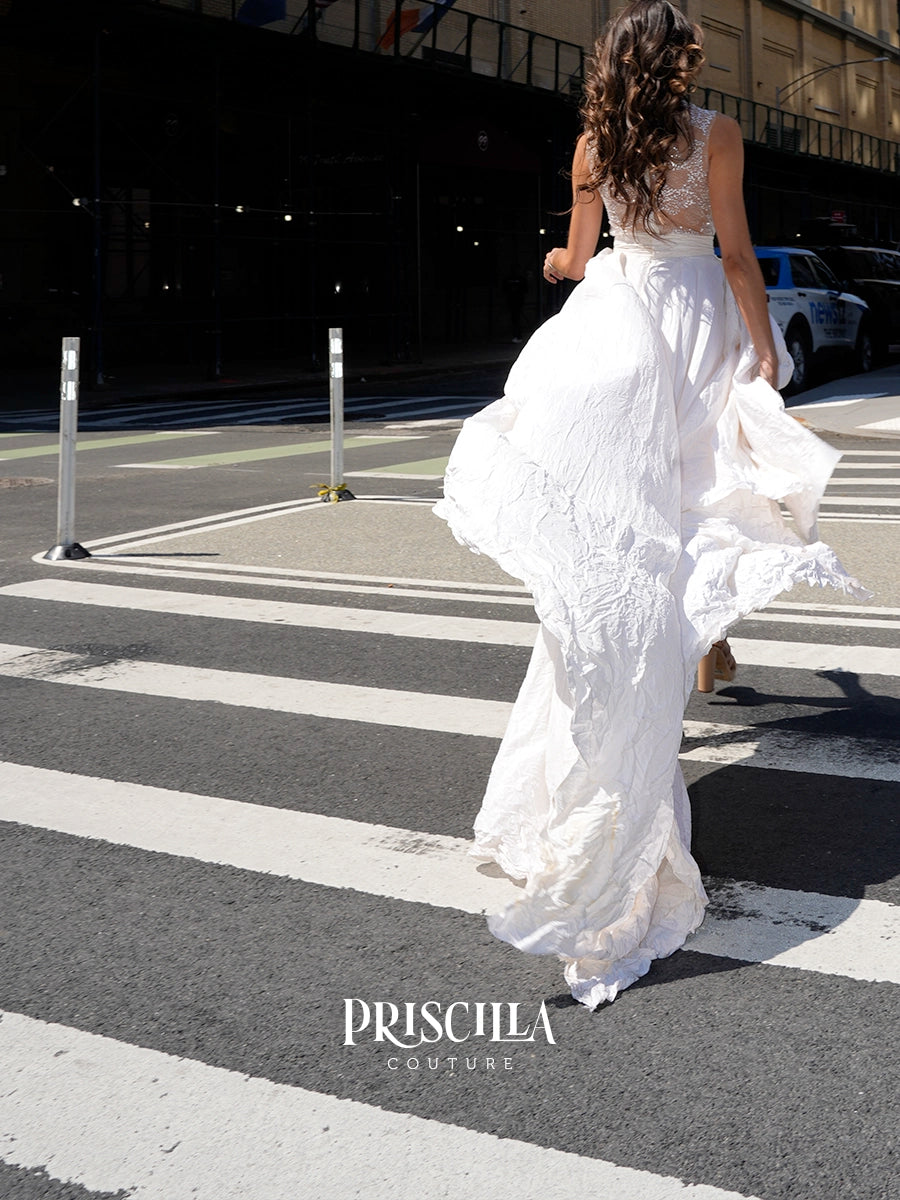 A woman wearing a white Priscilla Couture sleeveless maxi dress walks across a city crosswalk, viewed from behind as the dress billows in the wind.