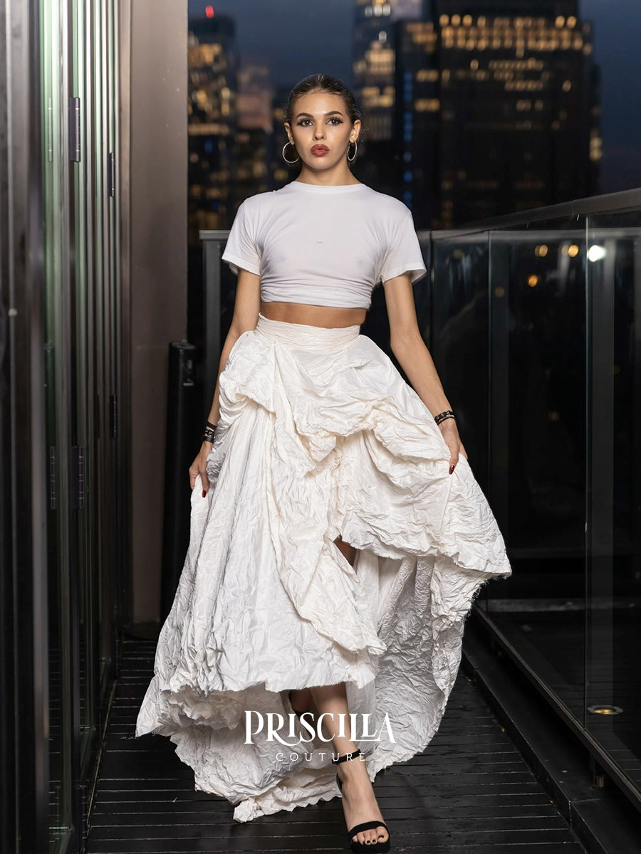 A woman stands on a balcony at night wearing a white short-sleeve crop top and a voluminous, heavily textured white ruffled skirt. The background shows a blurred city skyline at dusk.