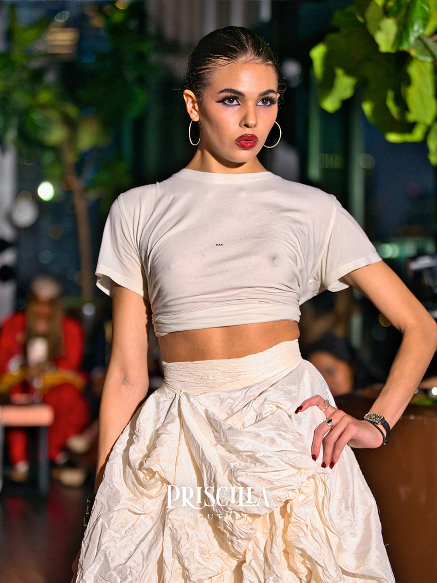 A runway model wearing a cream-colored two-piece outfit consisting of a short-sleeve crop top and a textured, voluminous skirt. The image is taken on a fashion runway with a blurred background of plants and audience members.