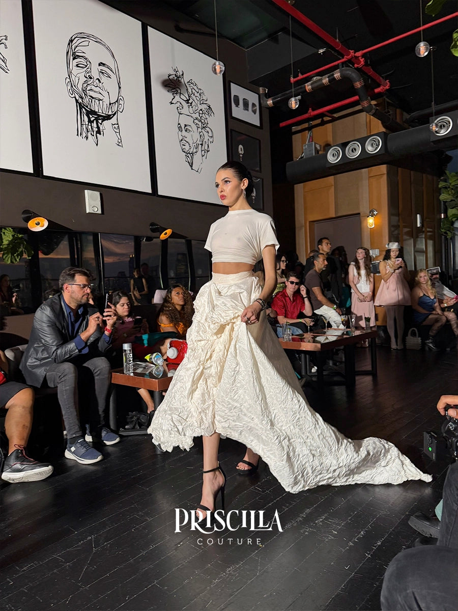 A model walks a fashion runway wearing a Priscilla Couture outfit consisting of a white short-sleeve crop top and a voluminous textured white skirt with a long train. The runway is held in a dark, stylish venue with an audience seated along the sides.