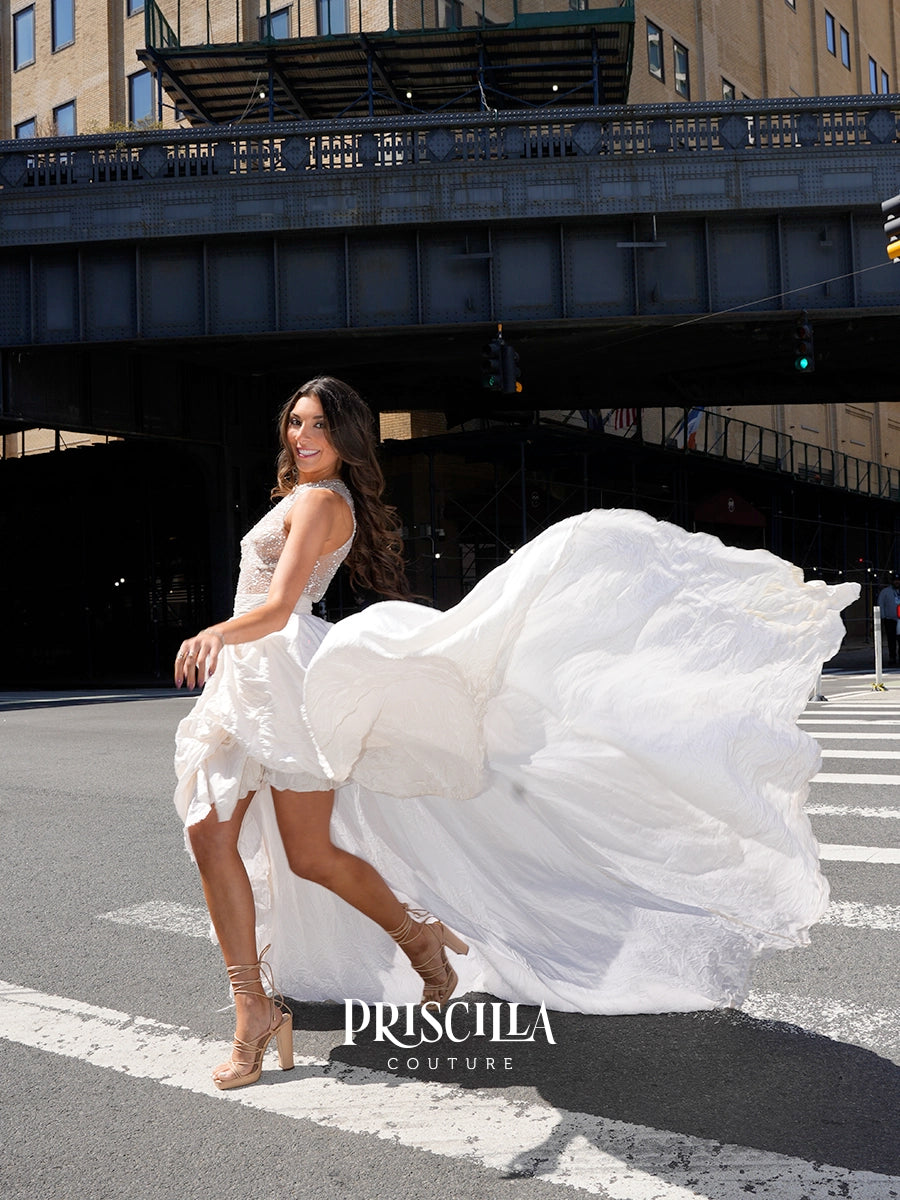 A woman in a white Priscilla Couture wedding dress with a dramatic flowing train poses on a city street under an elevated train track. The brand name 'PRISCILLA COUTURE' is displayed at the bottom.