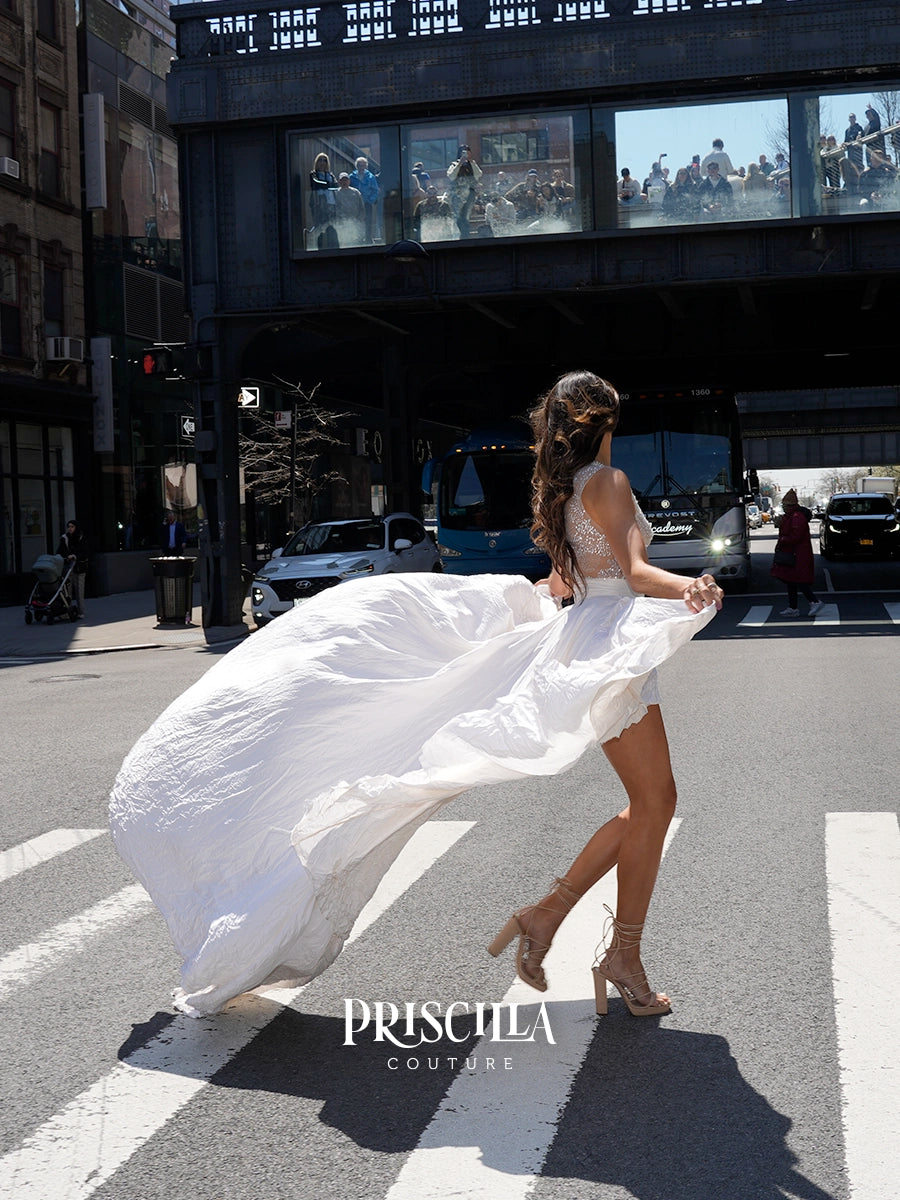 A woman in a white sleeveless dress with a flowing sheer skirt walks across a city crosswalk under an elevated train track. The brand name 'PRISCILLA COUTURE' is displayed at the bottom of the image.
