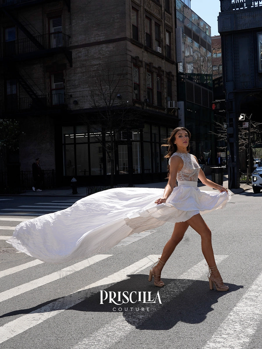 A woman walks across a city street crosswalk wearing a white PriscilLa Couture mini dress with a sheer lace bodice and a flowing white skirt that billows in the wind. 