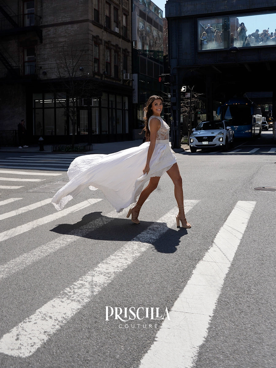 A woman in a flowing white sleeveless dress walks across a city crosswalk, with buildings, cars, and an elevated track visible in the urban background. The brand name PRISCILLA COUTURE appears at the bottom.