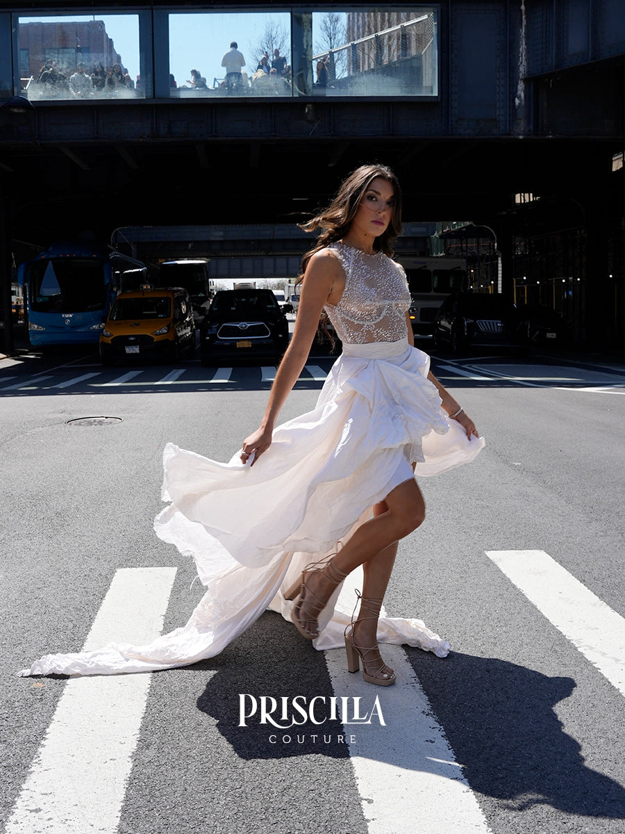 A woman modeling a white sleeveless gown with a sheer lace bodice and flowing skirt, standing on a city crosswalk under an elevated train track. The brand name 'PRISCILLA COUTURE' is watermarked at the bottom.