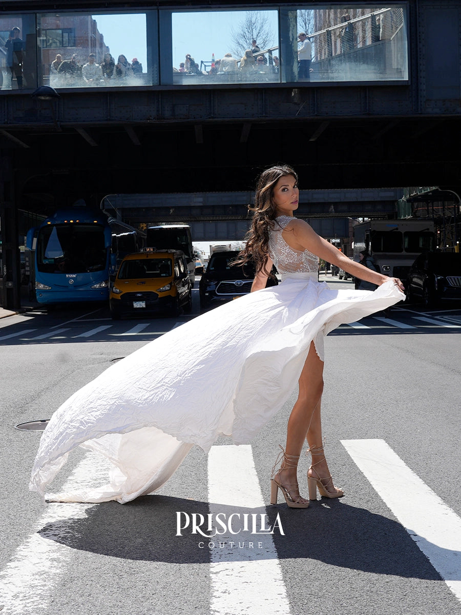 A woman in a white lace and taffeta wedding dress with a flowing train stands on a city crosswalk in front of an underpass, holding out one side of the dress. The brand name 'PRISCILLA COUTURE' is displayed at the bottom.