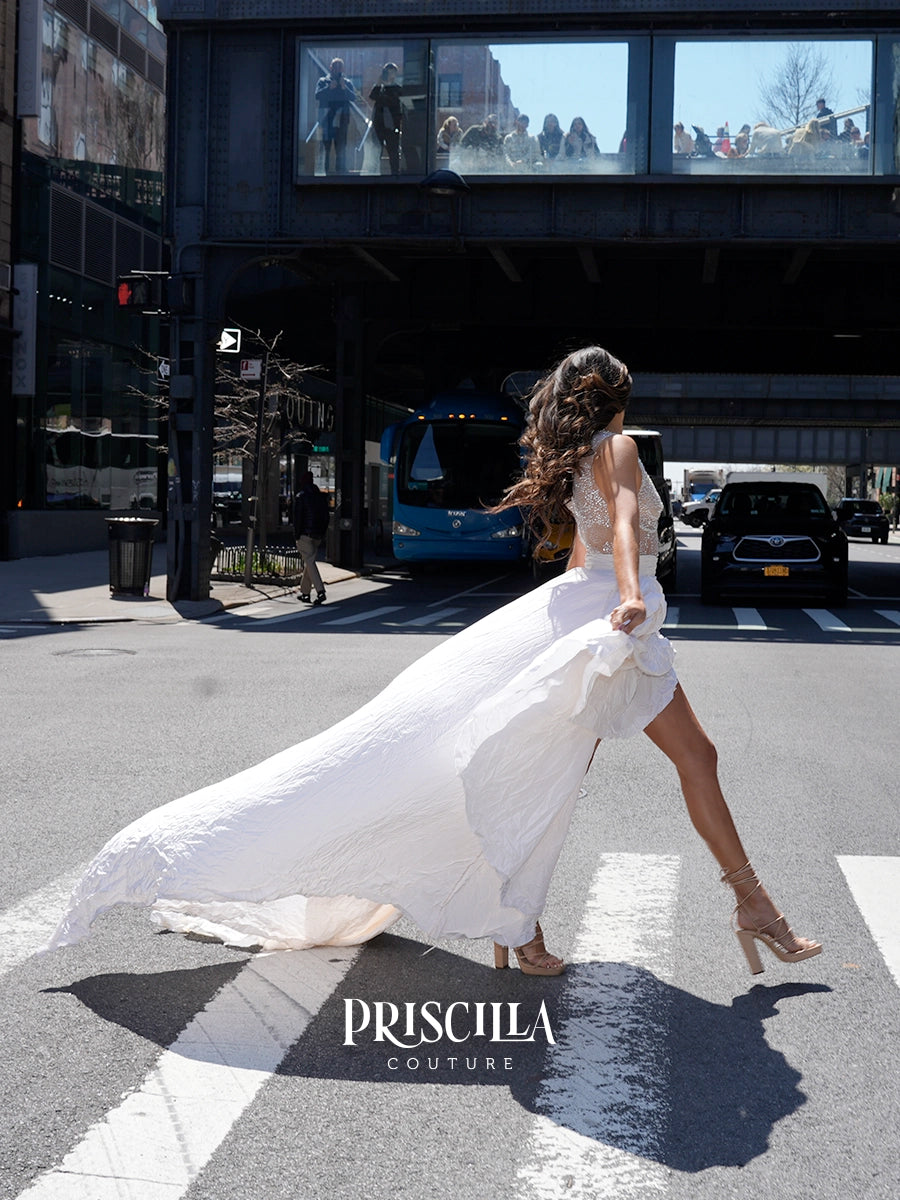 A woman in a flowing white Priscilla Couture taffeta wedding gown walks across a city crosswalk under an elevated train track, with a blue train and urban background visible behind her.