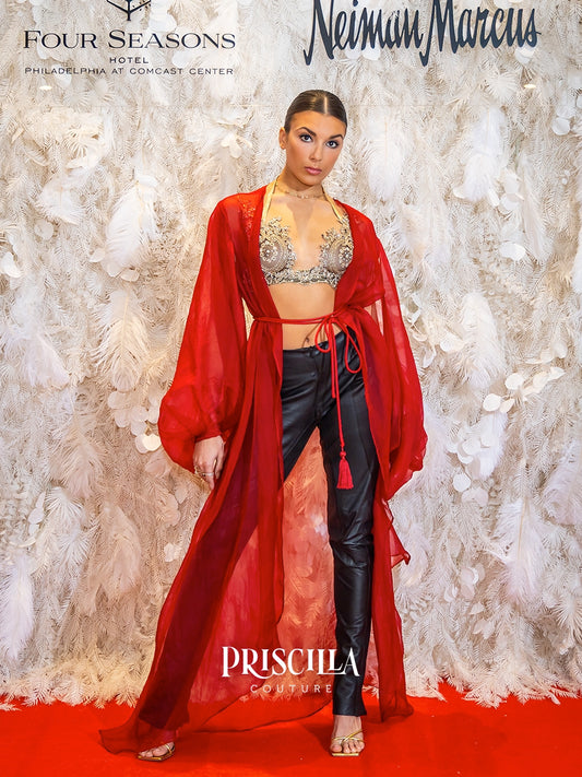 A model stands on a red carpet in front of a white feather backdrop, wearing a sheer red robe over a beige embellished bralette and black faux-leather pants. Visible text includes Four Seasons Hotel, Neiman Marcus, and Priscilla Couture.