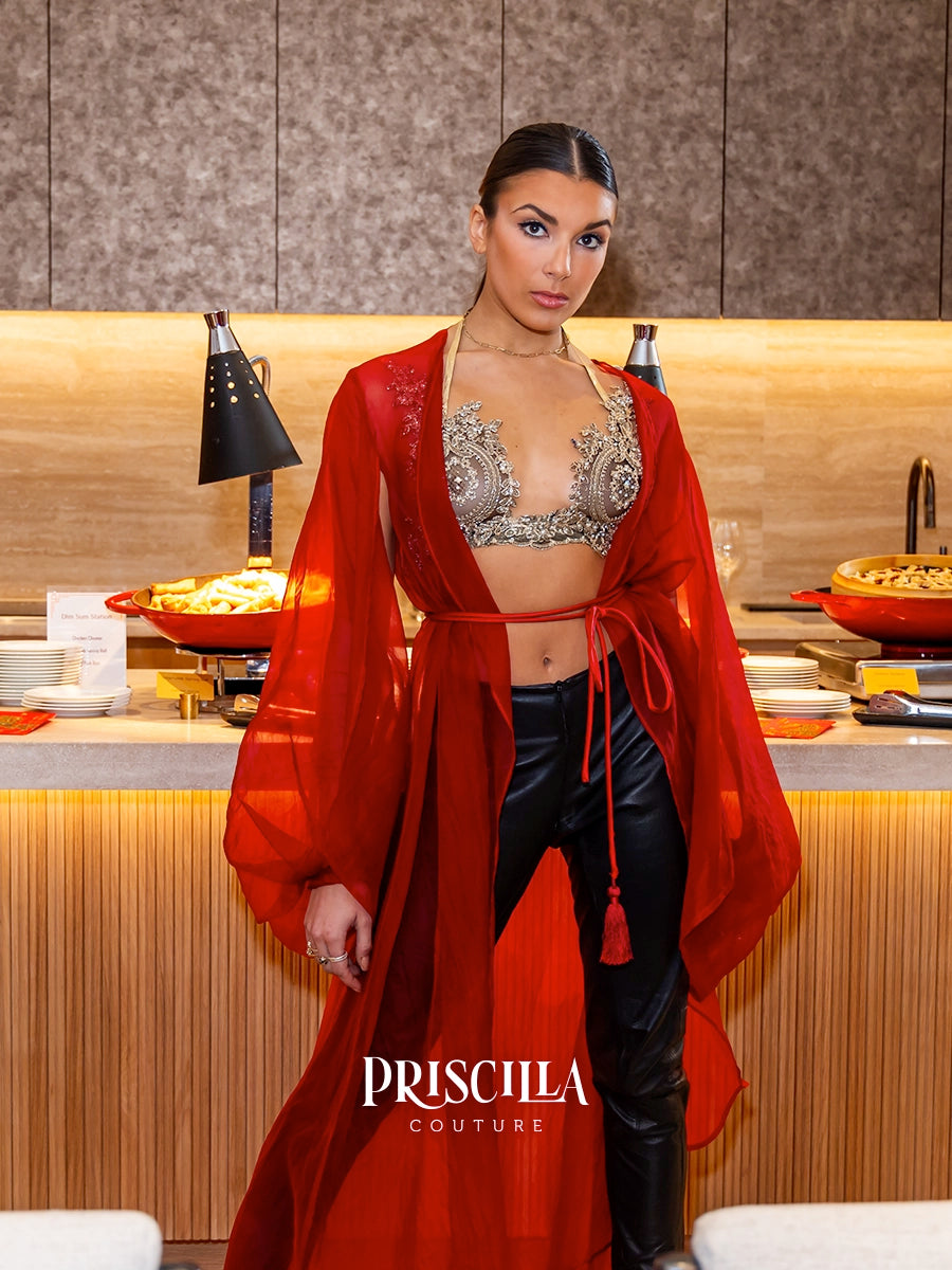 A woman models a red sheer open-front silk robe over a silver embellished bralette and black pants in a warm indoor setting with buffet tables in the background. The brand name 'PRISCILLA COUTURE' is displayed at the bottom.