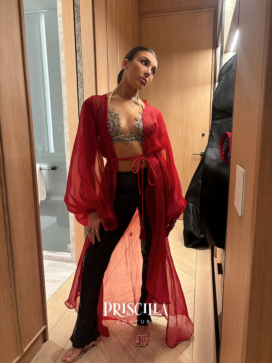 A woman models a red sheer long-sleeve wrap robe over a black pantsuit, standing in a hallway with wood-paneled walls. The brand name 'PRISCILLA COUTURE' is watermarked at the bottom.