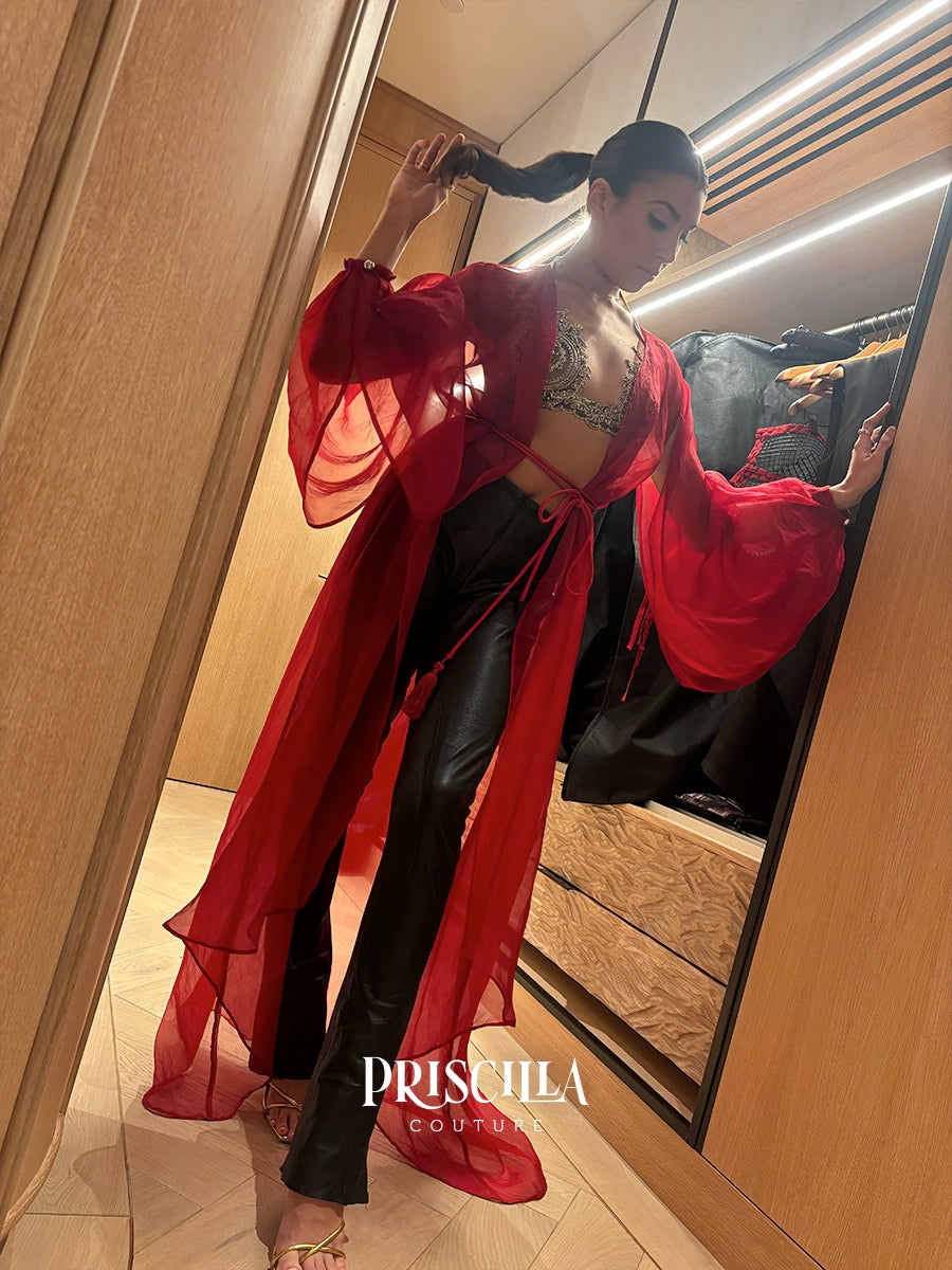 A woman in a fitting room wearing a sheer red long-sleeve cover-up over a green lace bralette and black pants, with a mirror reflecting her profile. The brand name 'PRISCILLA COUTURE' is watermarked at the bottom.