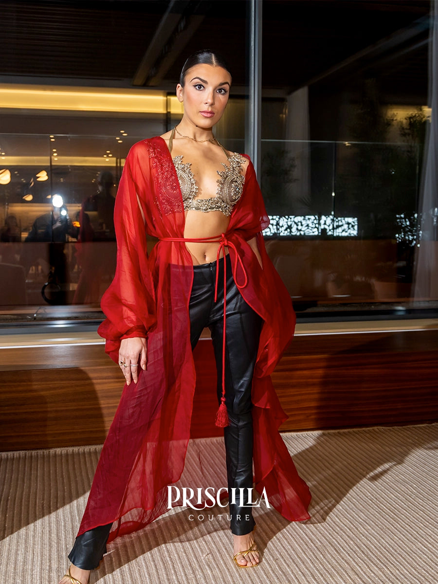 A woman models a red sheer long-sleeve silk drape cover-up with a matching waist tie, worn over a beaded bralette and black pants, in an indoor setting with a glass window background. The brand name 'PRISCILLA COUTURE' is watermarked at the bottom.