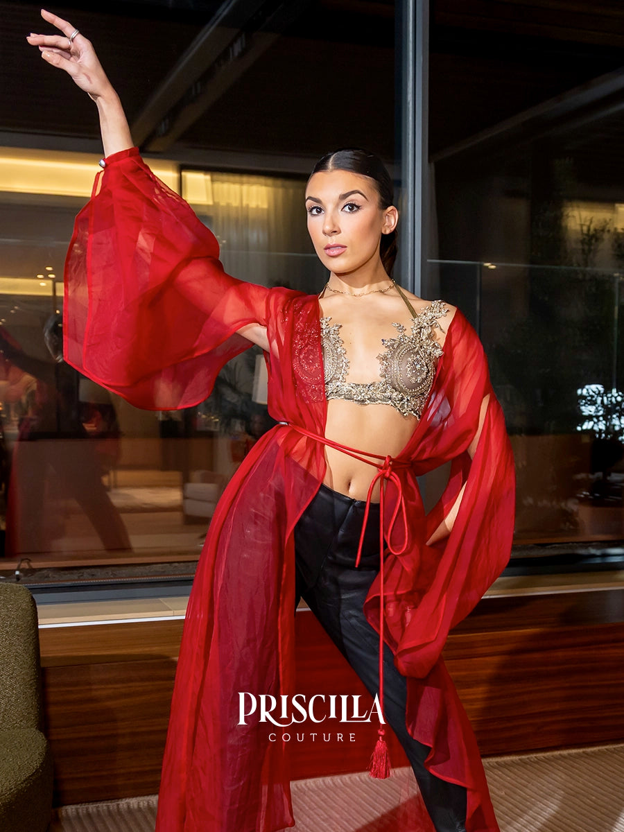 A model poses in a red sheer long-sleeve silk organza robe tied at the waist over a lace-trimmed crop top and black pants, with the Priscilla Couture logo visible at the bottom.
