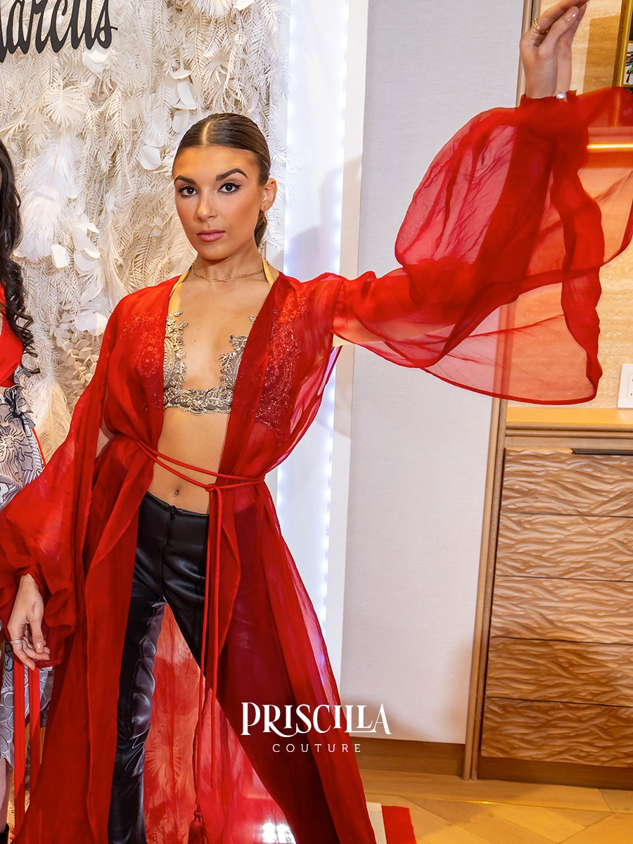 A model wearing a vibrant red sheer long-sleeve silk robe over a silver bralette and black pants, posing indoors with one arm raised. The brand name 'PRISCILLA COUTURE' is visible as a watermark at the bottom.