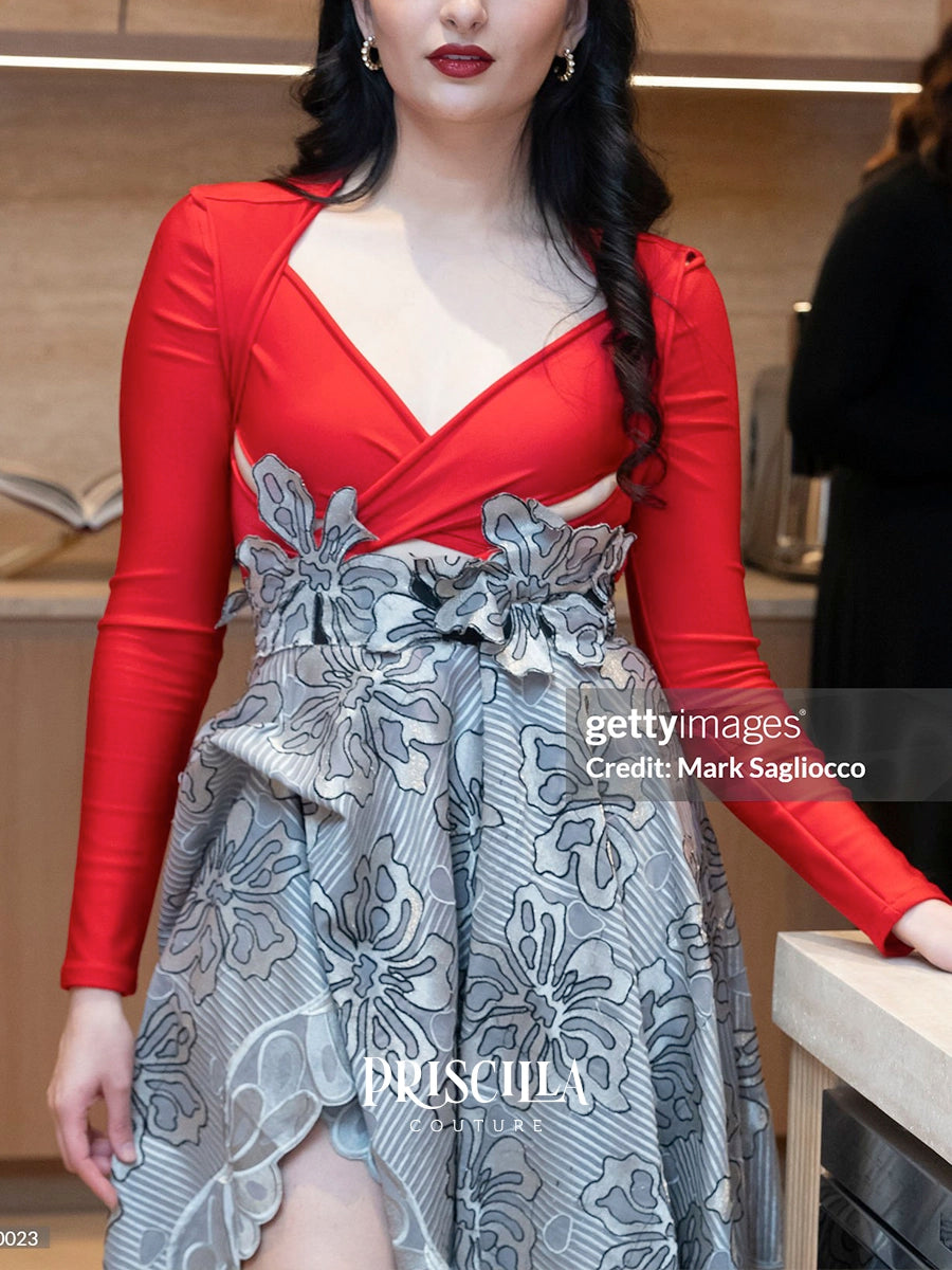 A woman wearing a red long-sleeve wrap-style top with a gray and white floral-patterned skirt, standing indoors near a counter. A Getty Images watermark and the brand name PRISCILLA COUTURE are visible.