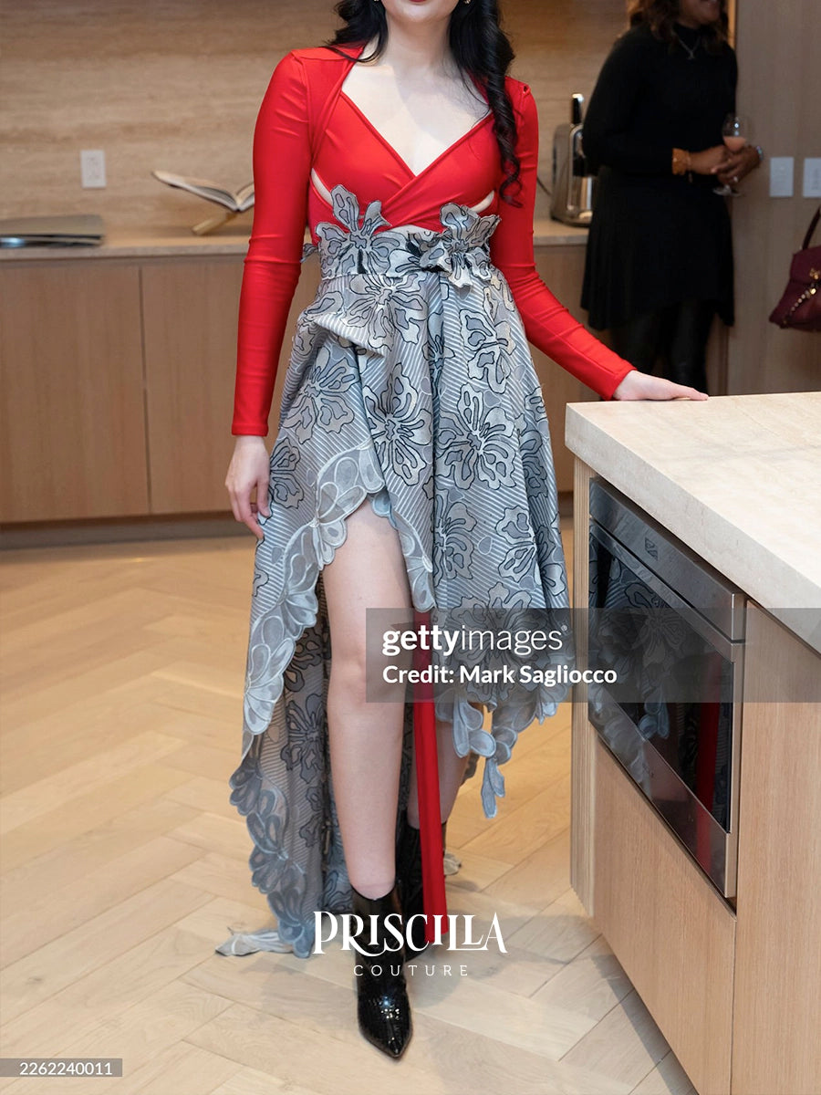 A woman models a Priscilla Couture dress in a modern kitchen setting. The dress features a vibrant red long-sleeve bodice with a sweetheart neckline and a high-low skirt made of a gray and white floral jacquard fabric with a matching waist bow.