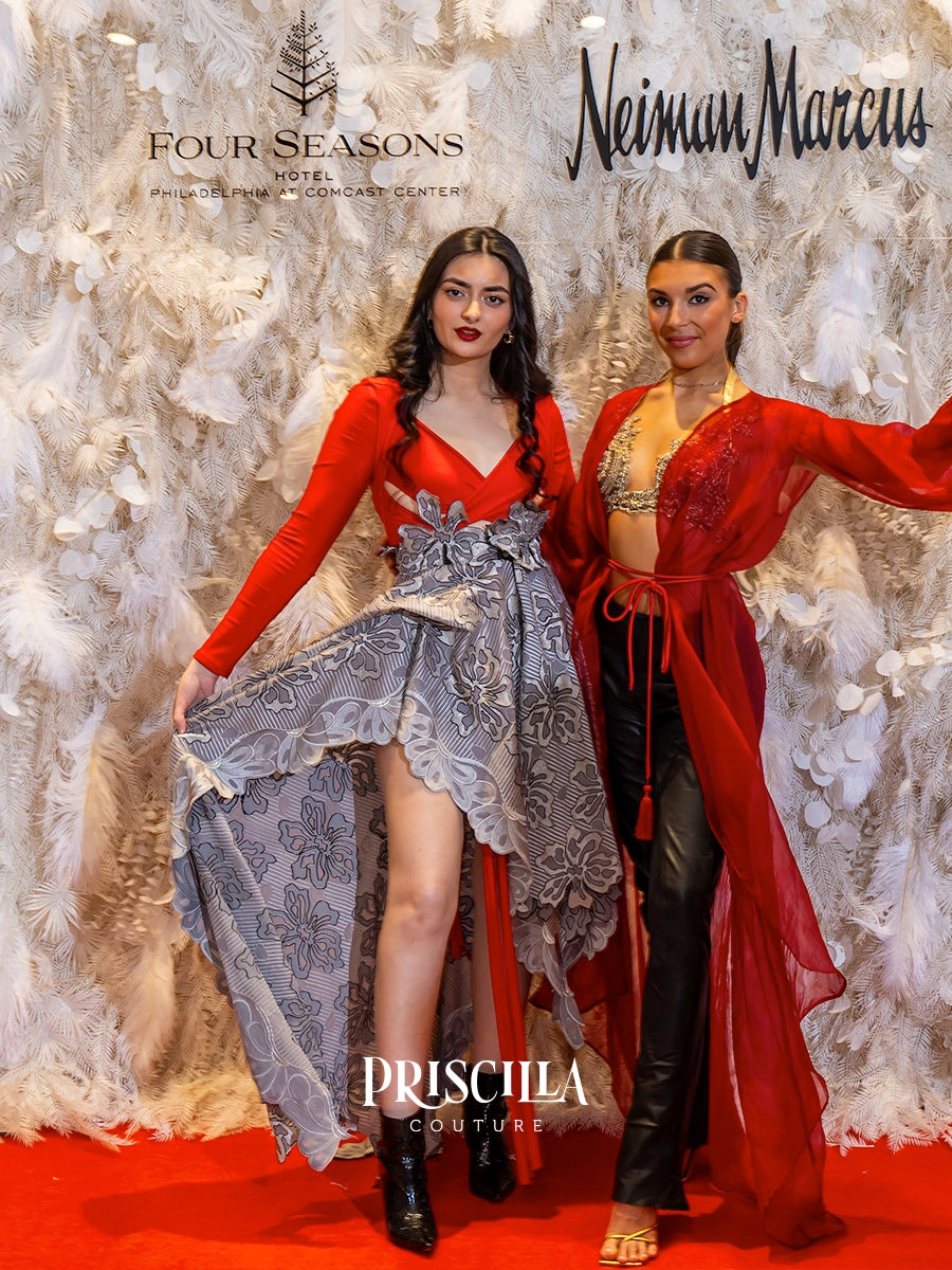 Two women pose in elaborate red and gray formal gowns in front of a white feathered backdrop. The left woman wears a red long-sleeve dress with a sheer gray floral skirt, while the right woman wears a matching red open-front robe over black pants.
