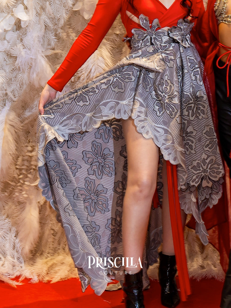 A close-up fashion photo of a model wearing a dramatic gray floral high-low skirt with a red long-sleeve top, holding up the skirt to show the leg slit. The brand name 'PRISCILLA COUTURE' is watermarked at the bottom.