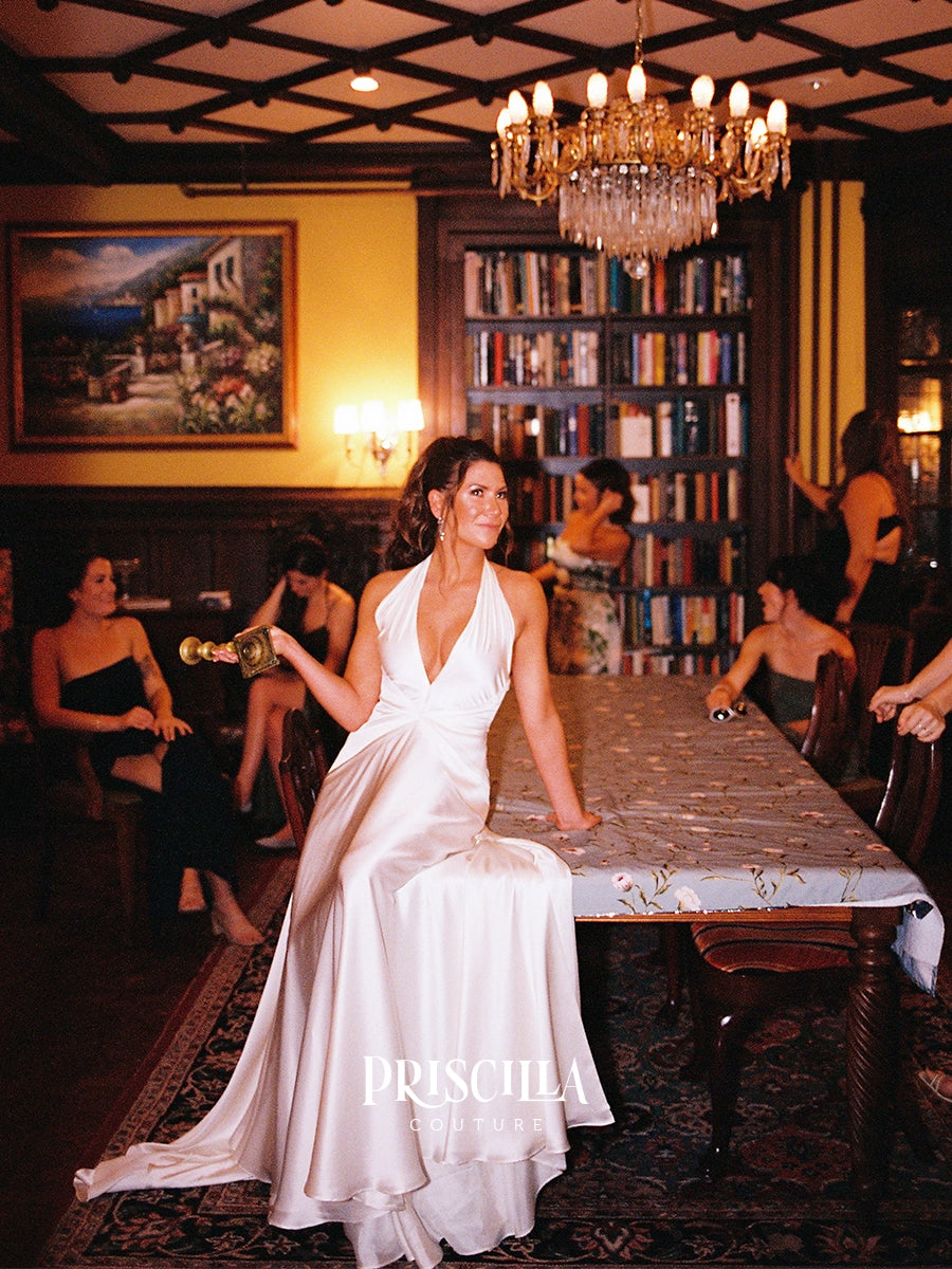 A bride in a Priscilla Couture custom white satin halter gown sits at a long table in a warmly lit indoor venue with bookshelves, chandeliers, and other guests visible in the background. The brand name PRISCILLA COUTURE is printed in white text at the bottom center.