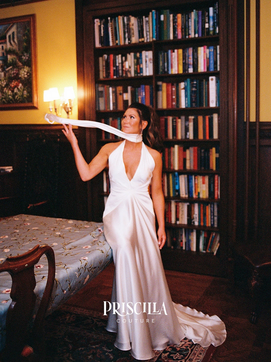 A bride in a Priscilla Couture custom white halter-neck floor-length gown stands in a room with tall bookshelves, holding a long white ribbon or sash across the frame. The brand name PRISCILLA COUTURE appears at the bottom.