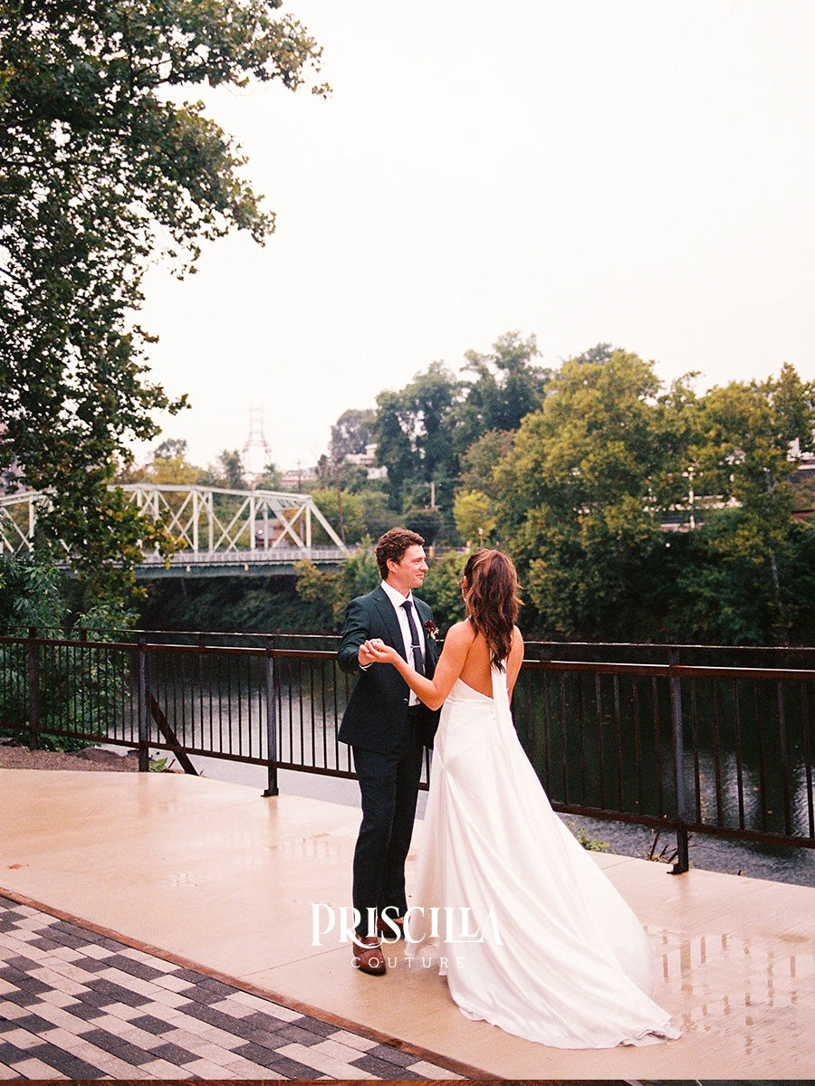 A bride in a Priscilla Couture custom sleeveless white wedding gown and a groom in a black suit pose together on an outdoor deck overlooking water and trees, with a bridge in the background. The name Priscilla appears as a watermark near the bottom.