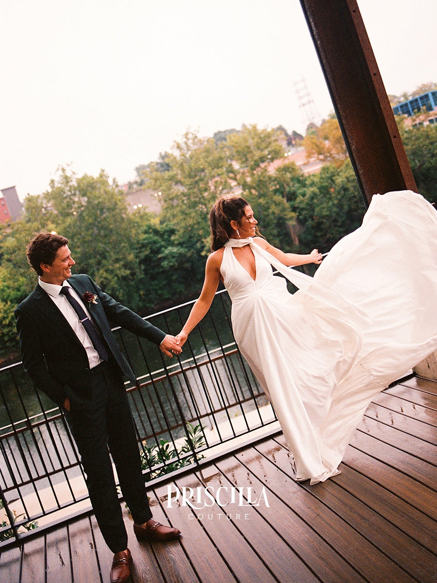 A bride in a Priscilla Couture custom white sleeveless wedding gown with a deep V-neck and a groom in a black suit holding hands on a wooden deck, with the bride's dress being blown in the wind. Trees and buildings are visible in the background.