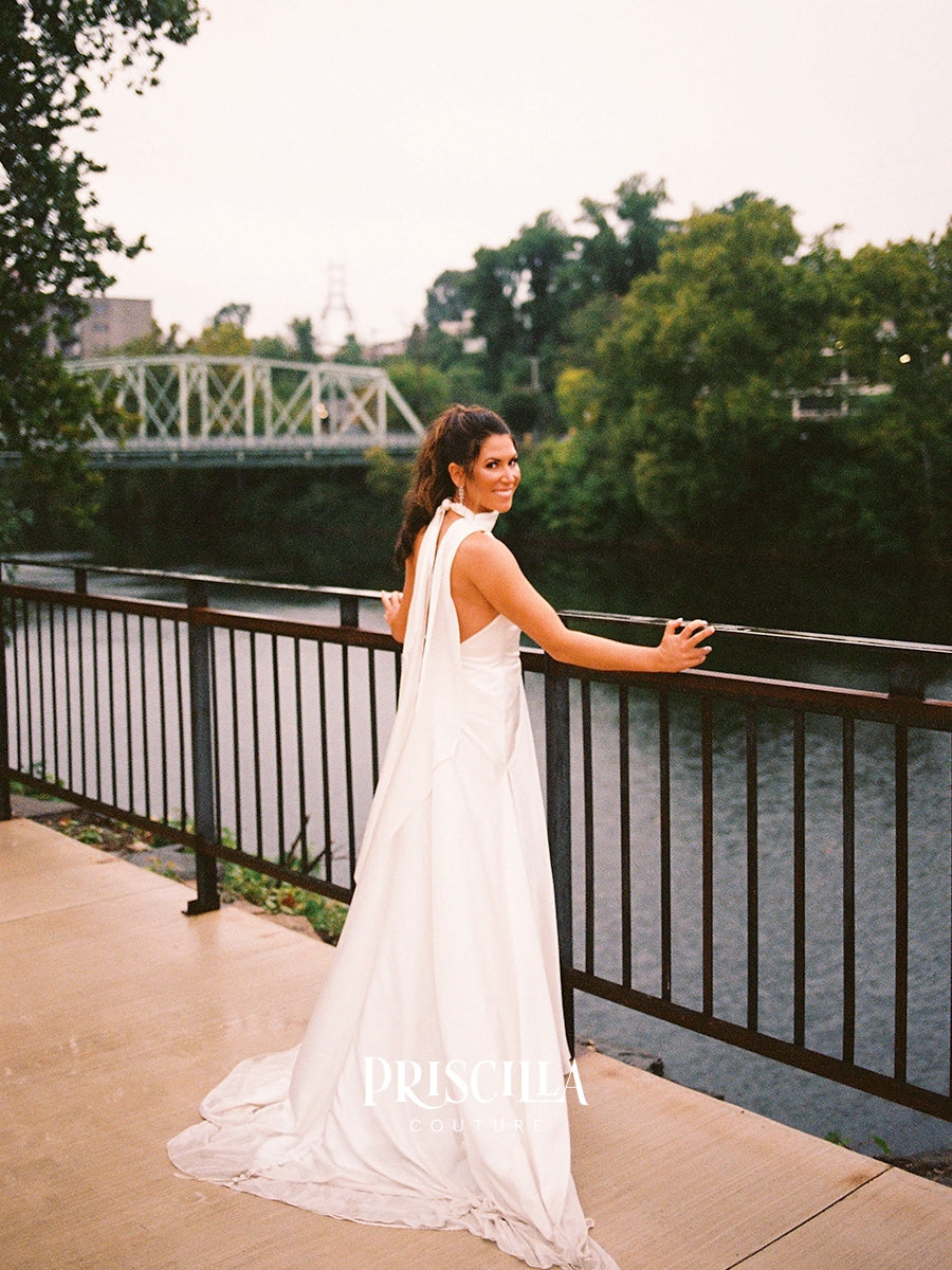 A bride in a Priscilla Couture custom  sleeveless white wedding gown with a high neck and back stands on a riverside walkway beside a railing, looking over the water. Trees, water, and a bridge are visible in the background.
