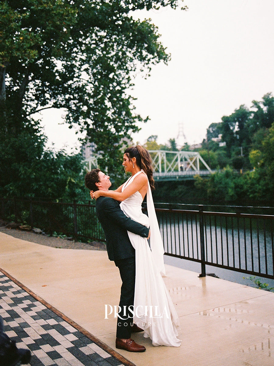 A bride in a Priscilla Couture custom white wedding gown and veil and a groom in a dark suit share an intimate moment, being lifted by the groom on a riverside walkway with trees, water, and a bridge in the background. The brand name 'PRISCILLA COUTURE' is watermarked at the bottom.