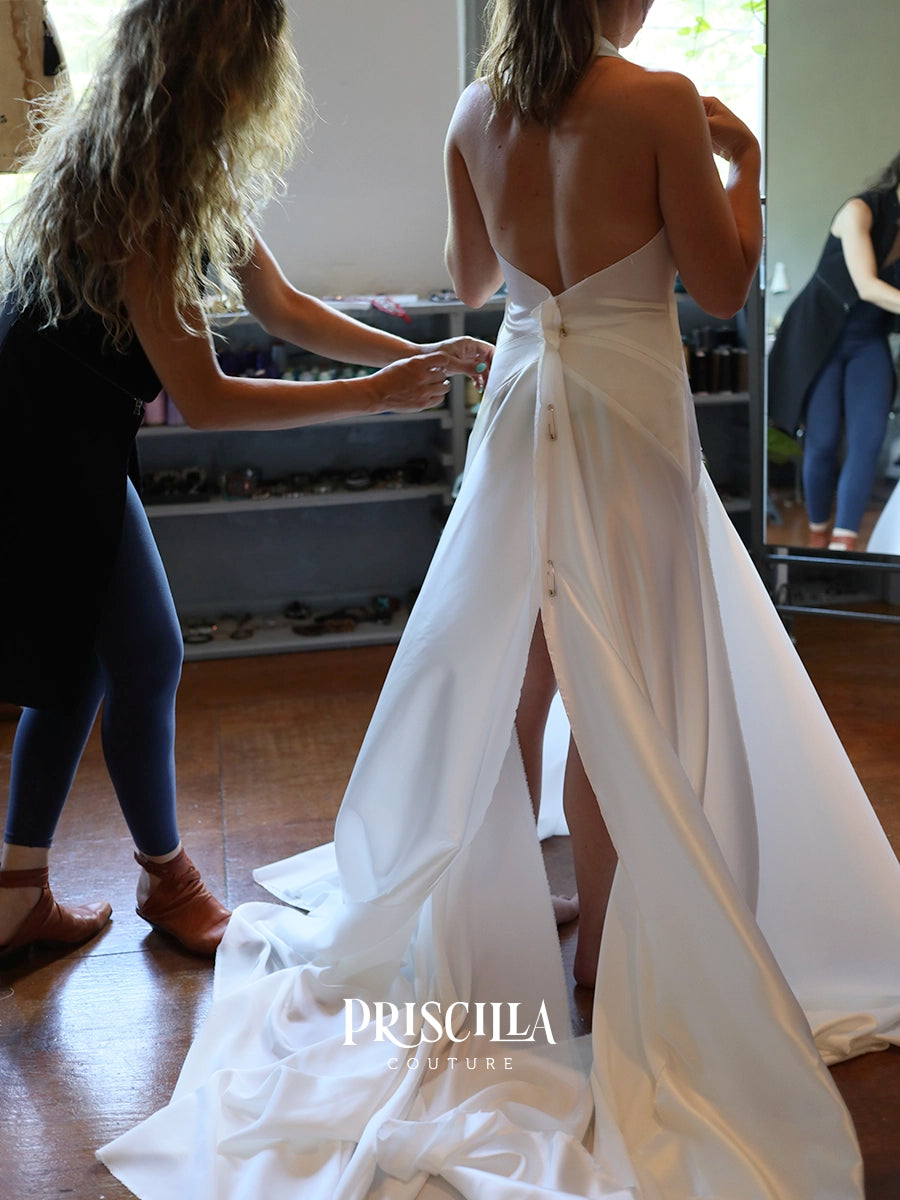 A bride at a fitting session wearing a Priscilla Couture custom white satin backless gown with a large bow at the back is having her hair styled by a fashion designer in a room with wooden floors and a mirror. The brand name 'PRISCILLA COUTURE' is watermarked at the bottom.
