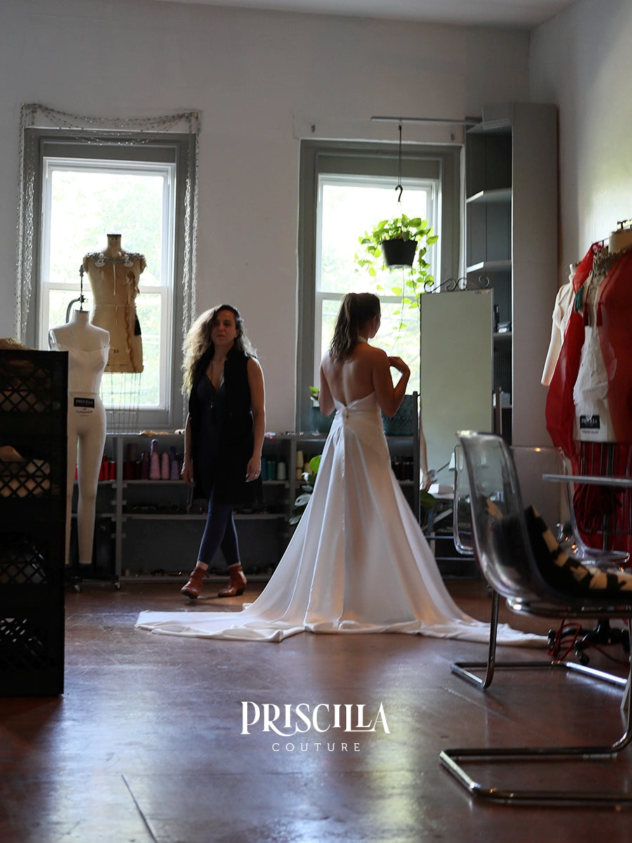 A bride in a fitting session wearing a Priscilla Couture custom white wedding gown stands with her back to the camera in a bridal salon or dressing room, with another woman nearby and mannequins and clothing racks visible in the room. The brand name 'PRISCILLA COUTURE' is displayed at the bottom.